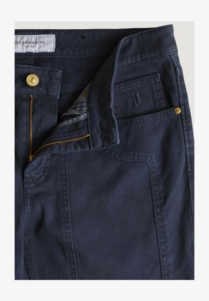 Granatowe spodnie z częściowo rozpiętym złotym zamkiem błyskawicznym, zapięciem na guzik, przednimi kieszeniami i białą metką z napisem „Jeckerson EST. 1995” wewnątrz pasa.