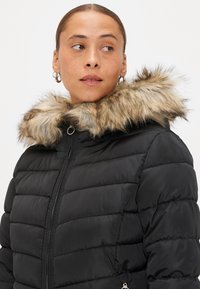 Casaco puffer preto com capuz forrado de pele, apresentando um design acolchoado, fecho de correr na frente e bolsos laterais. A pele tem um padrão castanho claro.