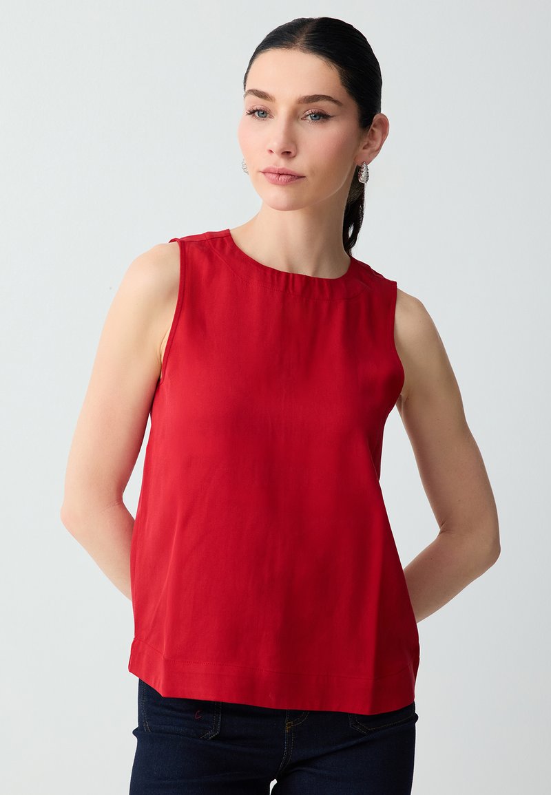 Femme aux cheveux foncés tirés en arrière, portant des boucles d’oreilles argentées en forme de créoles et un haut rouge sans manches sur un fond gris clair uni.