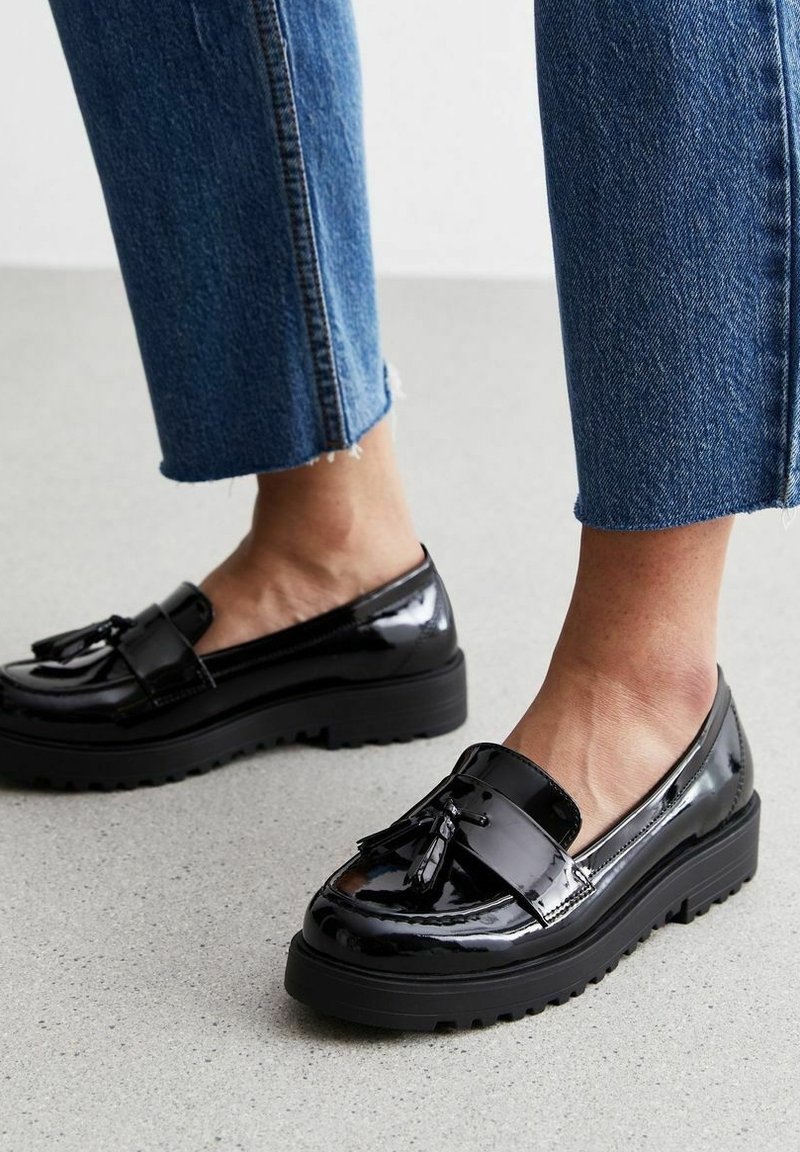 Pieds portant des mocassins noirs à plateforme brillante avec des glands, associés à un jean en denim bleu raccourci sur un sol gris clair.