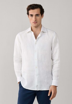 Hombre con camisa blanca de manga larga abotonada y pantalones azul oscuro, de pie frente a un fondo gris liso.