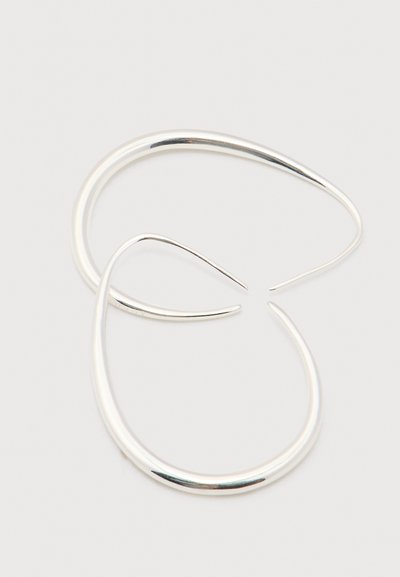 Dwa gładkie, srebrne kolczyki w otwartą obręcz z zwężającymi się końcami na jasnym tle.