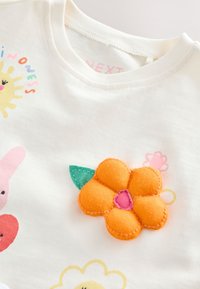 White cotton t-shirt featuring colorful appliqué designs, including a prominent orange flower with pink accents and other playful motifs.