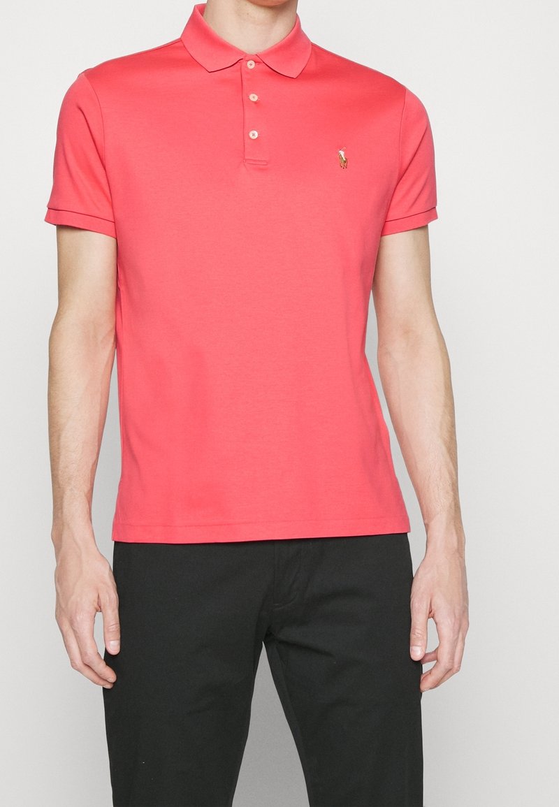 Homme portant un polo corail à manches courtes avec trois boutons et un pantalon noir, debout devant un fond clair uni.
