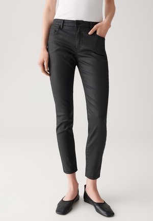 Femme portant un pantalon noir slim avec poches avant et des chaussures plates noires, debout devant un fond clair uni.