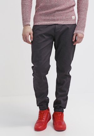 Mand iført mørke joggebukser, en lyserød tekstureret sweater og knaldrøde høje sneakers, stående på en hvid baggrund.