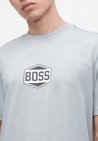 Νεαρό άτομο που φοράει γκρι ανοιχτό T-shirt με ένα ασπρόμαυρο εξαγωνικό λογότυπο που γράφει "BOSS" στο στήθος.