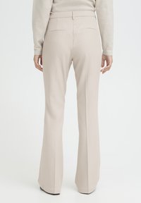 Personne portant un pantalon beige sur mesure et un haut à manches longues beige clair, vue de dos sur un fond blanc uni.