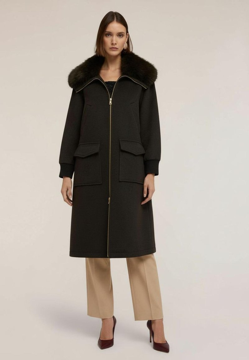 Lång svart kappa med pälskrage, guld dragkedja och två stora fickor. Bärs över beige byxor och burgundy klackar. Slät textur.