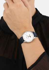 Am Handgelenk getragen, eine silberne Uhr mit weißem Ziffernblatt, blauem Lederarmband, Mondphasenanzeige und einem schwarzen Langarmkleidungsstück aus durchsichtigem Stoff.