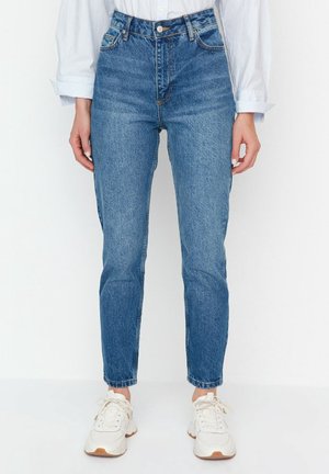 Persoon draagt hoog getailleerde blauwe spijkerbroek, witte sneakers en een wit shirt met lange mouwen, staand tegen een effen achtergrond.