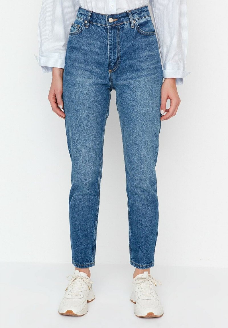 Personne portant un jean en denim bleu taille haute, des baskets blanches et une chemise blanche à manches longues, debout devant un fond uni.