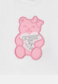 Material alb cu un aplicat în formă de ursuleț de pluș roz, având un inimioară pe care scrie "GUESS USA?" brodat cu fir argintiu și un fundiță pe ureche.
