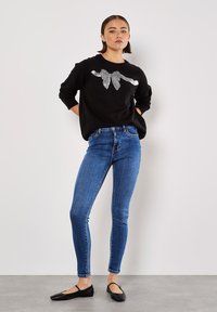 Maglione nero lavorato a maglia con dettaglio in pizzo argento, abbinato a jeans skinny blu. La modella indossa scarpe flat nere, in piedi con le mani sui fianchi.