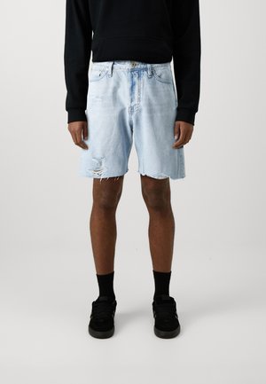 Helle blaue Jeansshorts mit einem fransigen Saum und distressed Details. Getragen mit schwarzen Socken und schwarzen Sneakers, kombiniert mit einem schwarzen Sweatshirt.
