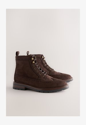 Brūnas zamšādas potīšu zābaki ar brogū detaļām, apaļu purngalu, metāla acu lūkām un teksturētu gumijas zolīti, ar nelielu papēdi un cilpiņu zābaku uzvilkšanai.