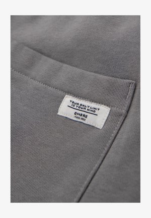 Tissu en coton gris avec une surface texturée. Étiquette blanche avec texte noir : "VOTRE SEUL LIMIT EST VOTRE ESPRIT OMBRE depuis 1985."