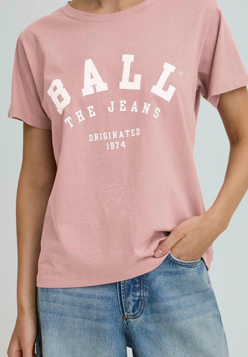 Osoba oblečená v ružovom tričku s nápisom "BALL THE JEANS ORIGINATED 1974" a modrých džínoch, s jednou rukou v vrecku.