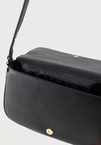 Μαύρη δερμάτινη τσάντα ώμου με χρυσό κουμπί κλεισίματος, ελαφρώς ανοιχτή που αποκαλύπτει το μαύρο εσωτερικό ύφασμα, και ρυθμιζόμενο λουρί με χρυσά μεταλλικά στοιχεία.