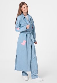 Manteau trench en denim bleu clair avec taille à nouer, présente des accents brodés roses et des poches. Ouverture à l'avant, manches longues et fentes latérales.