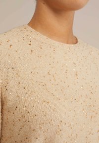 Beige stickad tröja med rund hals, prydd med spridda gyllene paljetter för textur. Mjukt tyg med en lätt och luftig känsla.
