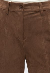 Gros plan sur un pantalon en daim marron avec fermeture à boutons et passants de ceinture, porté avec un haut côtelé blanc.