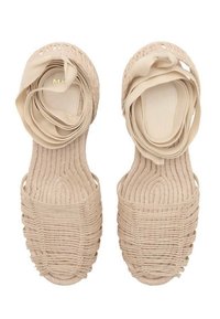 Beige woven sandals with an open toe design, featuring soft fabric straps that create a bow accent at the front. Textured surface and minimalist style.