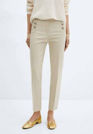 Pantalon beige slim avec des boutons dorés décoratifs à l'avant, porté avec un pull crème et des chaussures plates dorées sur un fond uni.