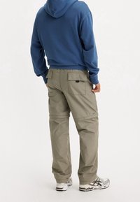 Khaki cargobroek met meerdere zakken, recht model en verstelbare manchetten, gecombineerd met een blauwe capuchontrui. Schoenen zijn grijs en wit.