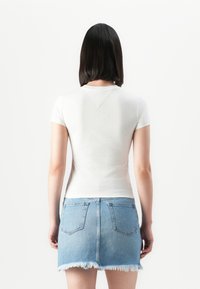 Femeie cu păr lung până la umeri, de culoare închisă, îmbrăcată cu o bluză albă cu mânecă scurtă și o fustă din denim albastru, ușor uzată, întoarsă cu fața spre un fundal simplu.