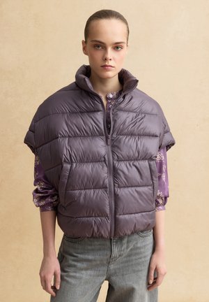 Junge Frau trägt eine ärmellose violette wattierte Jacke über einer geblümten langärmligen Bluse und hellgrauen Jeans, steht vor einem beigen Hintergrund.