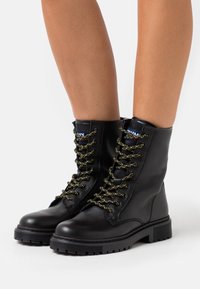 Botas de combate de cuero negro con cordones dorados y negros, punta redonda y suela de goma texturizada con un diseño de banda gruesa.