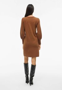 Robe en tricot marron avec de longues manches bouffantes, un col rond et des poignets côtelés, associée à des bottes noires montantes.