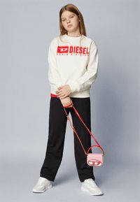Felpa bianca con logo rosso "DIESEL", pantaloni neri, sneakers bianche e una piccola borsa a tracolla rosa con dettagli e tracolla rossi.