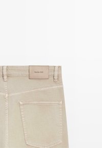 Beżowe spodnie dżinsowe z widoczną tylną kieszenią i szlufkami na pasek oraz skórzaną naszywką z napisem „Massimo Dutti” na talii.