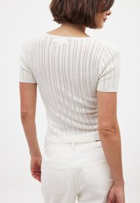 Hvid strikket top med vertikale striber, korte ærmer og en bølget udskæring. Stoffet har et tekstureret, åbent strikkemønster.