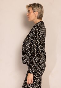 Schwarz-beiger Blazer mit geometrischem Muster, langen Ärmeln und zwei Fronttaschen, kombiniert mit passenden, taillierten Hosen.