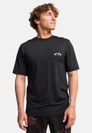 Mężczyzna ubrany w czarną koszulkę Billabong z krótkim rękawem oraz czarne szorty z czerwonym kwiatowym wzorem, spogląda na bok na białym tle.