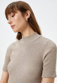 Top de punto acanalado beige con cuello alto y mangas cortas, que presenta un patrón texturizado y una silueta ajustada. Pendientes de aro dorados como acento.