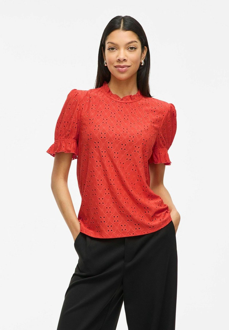Blusa in cotone rosso con motivo floreale in pizzo occhiello, maniche corte a palloncino e scollo scalloppato. Abbinata a pantaloni neri.