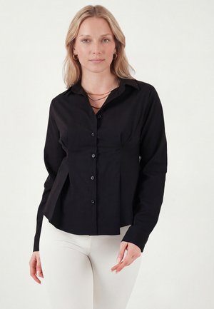 Blondynka ubrana w dopasowaną czarną koszulę na guziki z długimi rękawami i białe spodnie, stojąca na jednolitym tle.