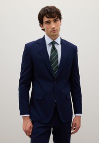 Completo blu navy con finitura texturizzata, design a uno petto e due tasche frontali. Indossato con una camicia azzurro chiaro e una cravatta a righe verdi.