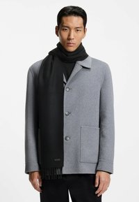 Cappotto di lana grigio con collo a punta e sciarpa di lana nera con dettagli frangiati. Il modello posa su uno sfondo bianco.