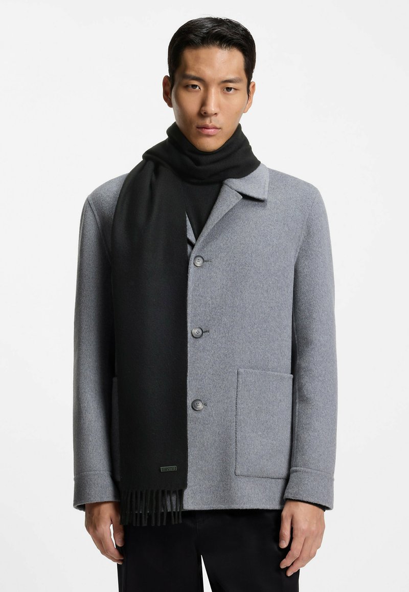 Cappotto di lana grigio con collo a punta e sciarpa di lana nera con dettagli frangiati. Il modello posa su uno sfondo bianco.