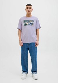 Světle fialové tričko s krátkým rukávem s tropickým grafickým potiskem a nápisem "JACK & JONES". Spárované s volnými modrými džíny a bílými teniskami.