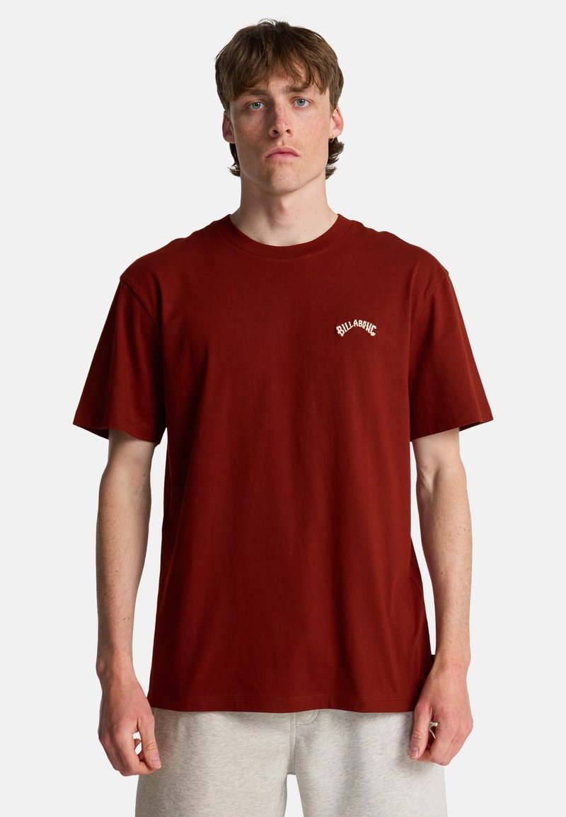 Junger Mann mit heller Haut und braunen Haaren trägt ein schlichtes dunkelrotes Billabong-T-Shirt und hellgraue Hosen, steht vor einem weißen Hintergrund.