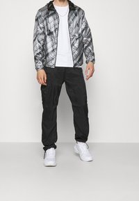 Sömsvart mönstrad vindjacka med huva, framfickor, som kombineras med svarta cargobyxor och vita sneakers. Slät, reflekterande yta.