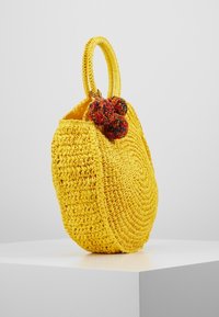 Okrągała, żółta torba pleciona z dwiema rączkami i kolorowymi pomponami zawieszonymi blisko górnej krawędzi.