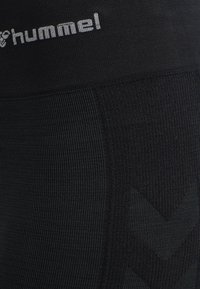 Zwarte sportlegging met een gestructureerde afwerking. Heeft een brede tailleband met het "hummel"-logo en een chevronpatroon in een iets donkerdere tint.