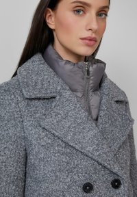Femme aux yeux bleus portant un manteau en laine gris texturé par-dessus une veste matelassée grise zippée, sur un fond clair uni.
